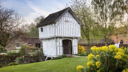 A view of the timber-framed Gatehouse at Brockhampton, surrounded by green lawns with yellow flowers blooming in the foreground, and other buildings in the background